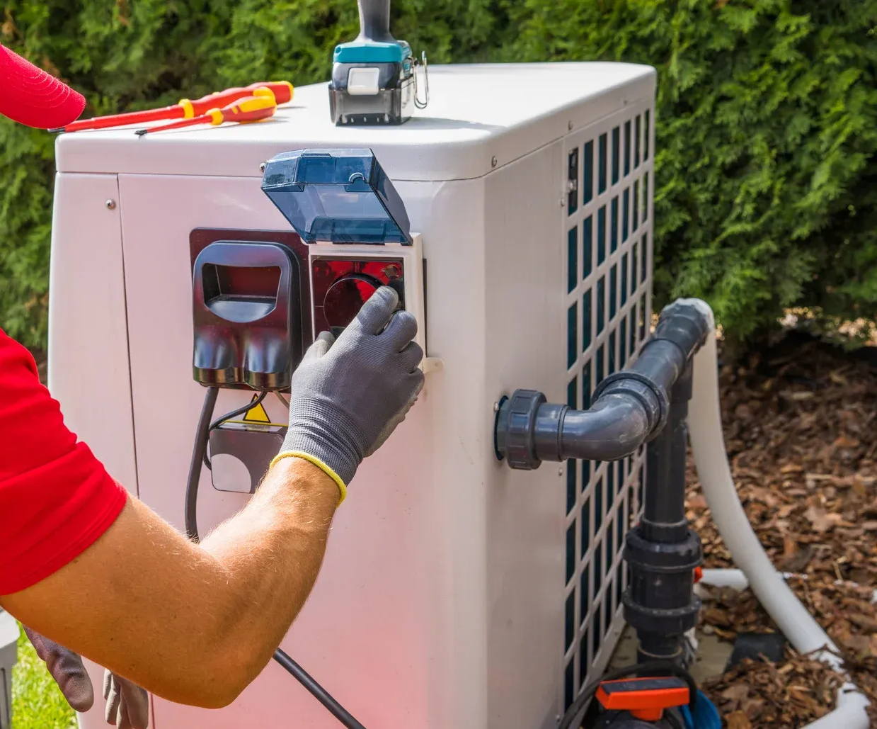 Technician adjusting controls on an outdoor air conditioning unit with exposed pipes and wiring