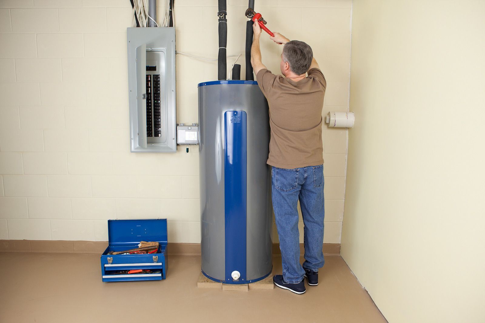Person inspecting electrical panel beside a water heater in a utility room, with tools on the floor