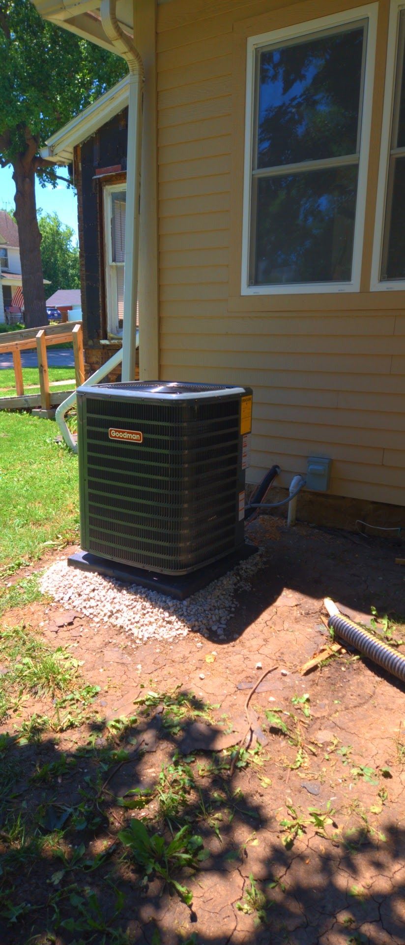 Outdoor air conditioner unit beside a tan house with a window and dirt yard