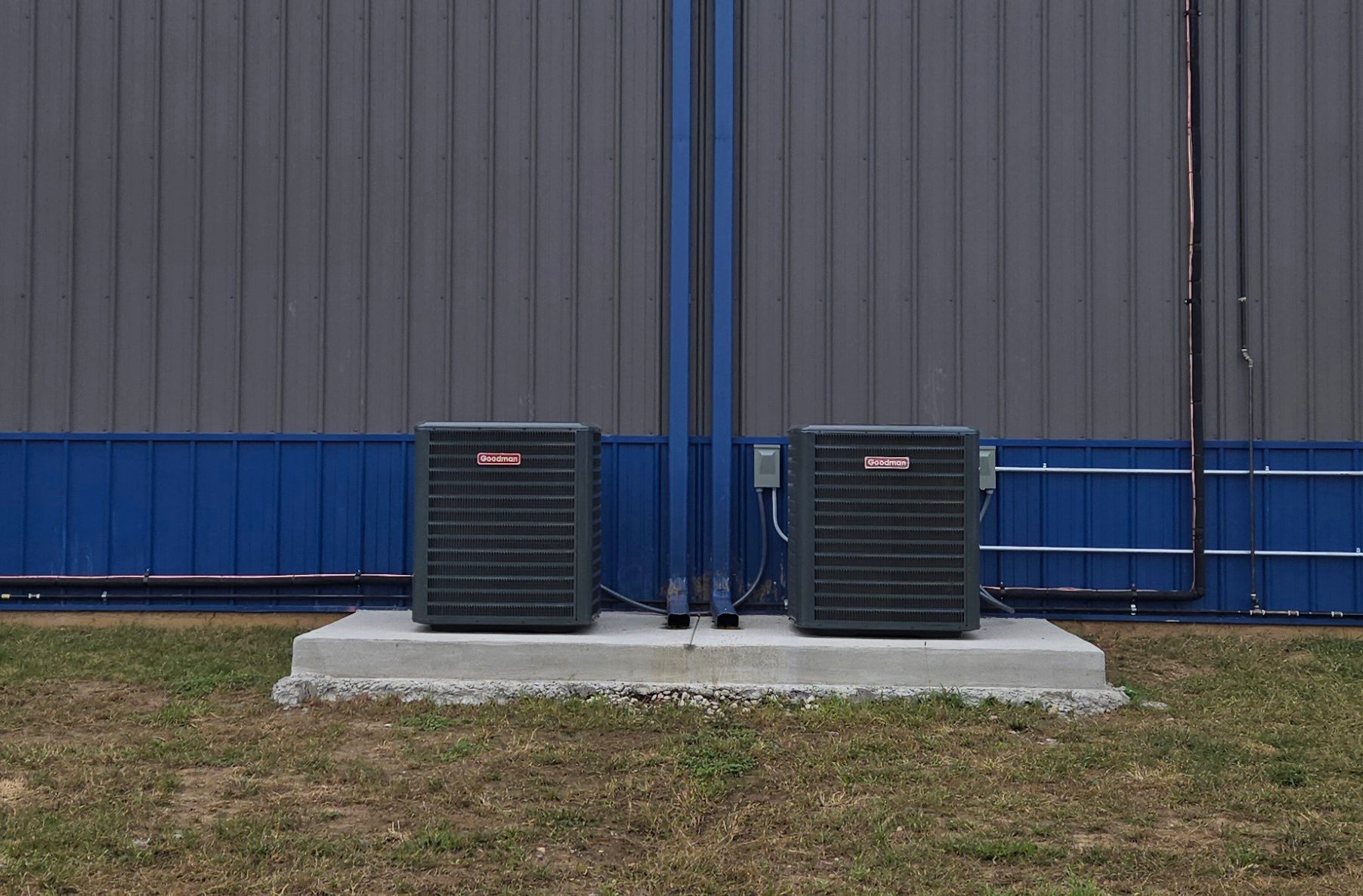 Two outdoor HVAC units on a concrete pad against a gray metal building with blue trim.
