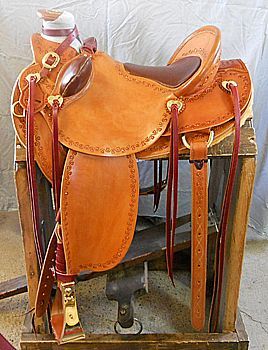 Tan leather western saddle on a wooden stand; details include dark brown seat, red accents, and gold hardware.