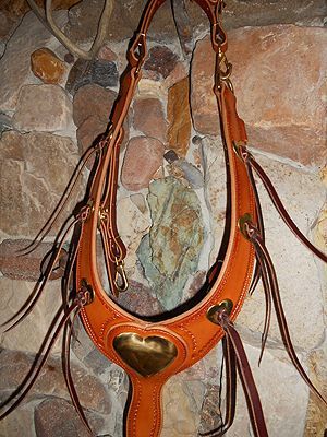 Brown leather horse breast collar with a heart-shaped metal plate, hanging on a stone wall.