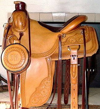 Tan leather western saddle with tooling, attached canteen, brown straps.