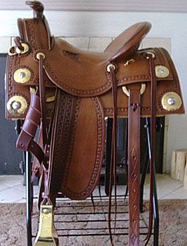 Brown leather western saddle with silver and gold accents, on a black stand indoors.