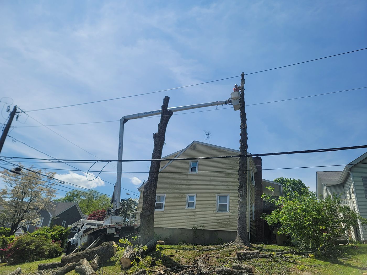 Tree trimming near a house with power lines. A truck with a lift arm supports a worker. Blue sky.