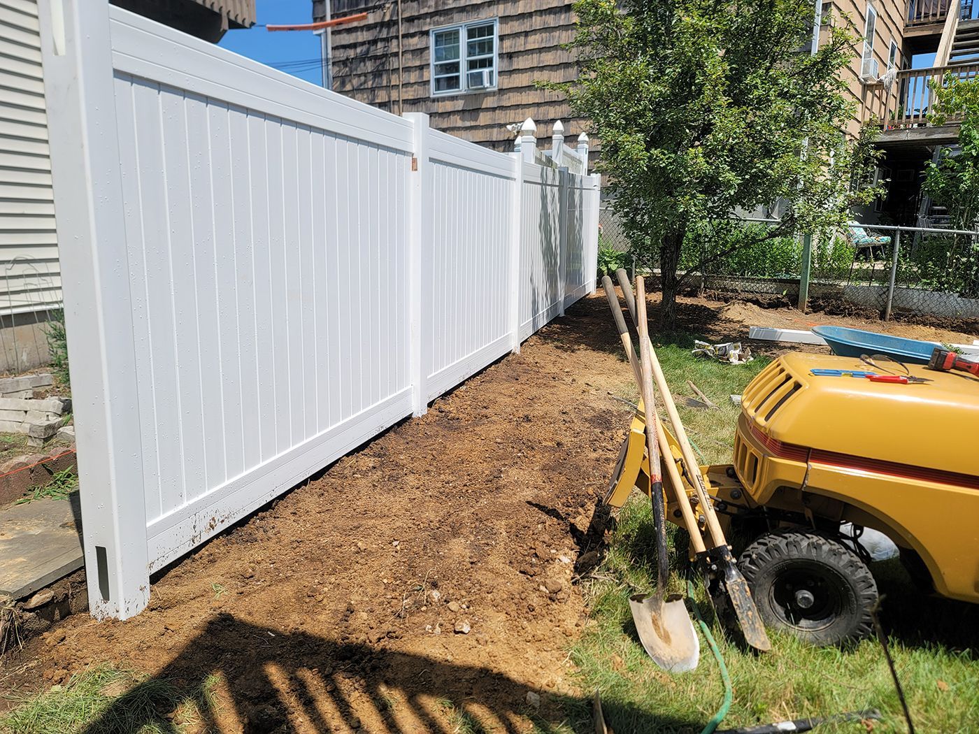 White vinyl fence in a yard with dirt and gardening tools next to a yellow utility vehicle.