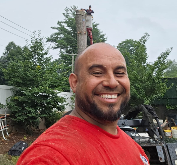 Smiling man in a red shirt outdoors, with trees and a utility pole in the background