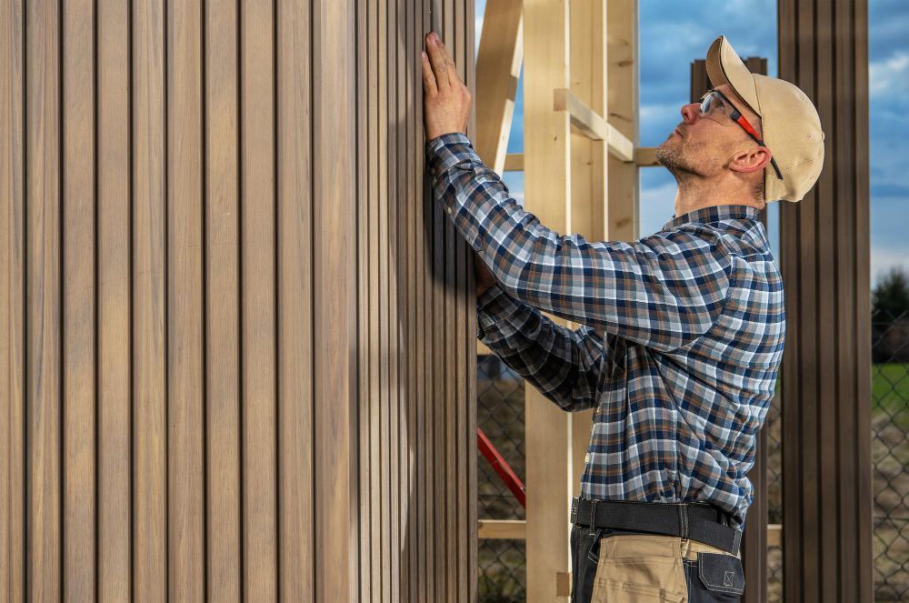 Worker in a cap installs vertical wooden slats on an exterior wall.