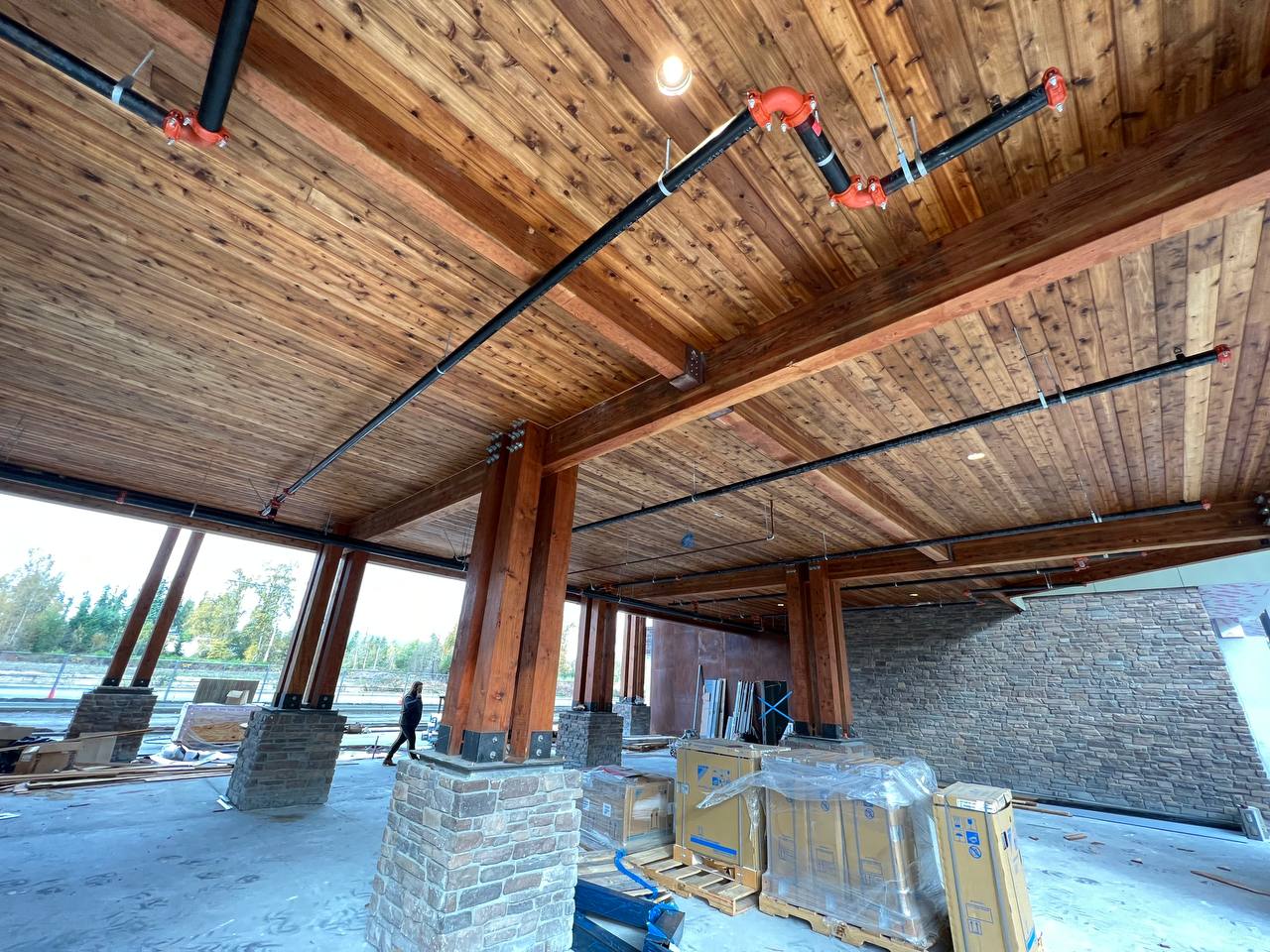 Open wooden pavilion under construction with brick columns, exposed ceiling beams, and wrapped materials.