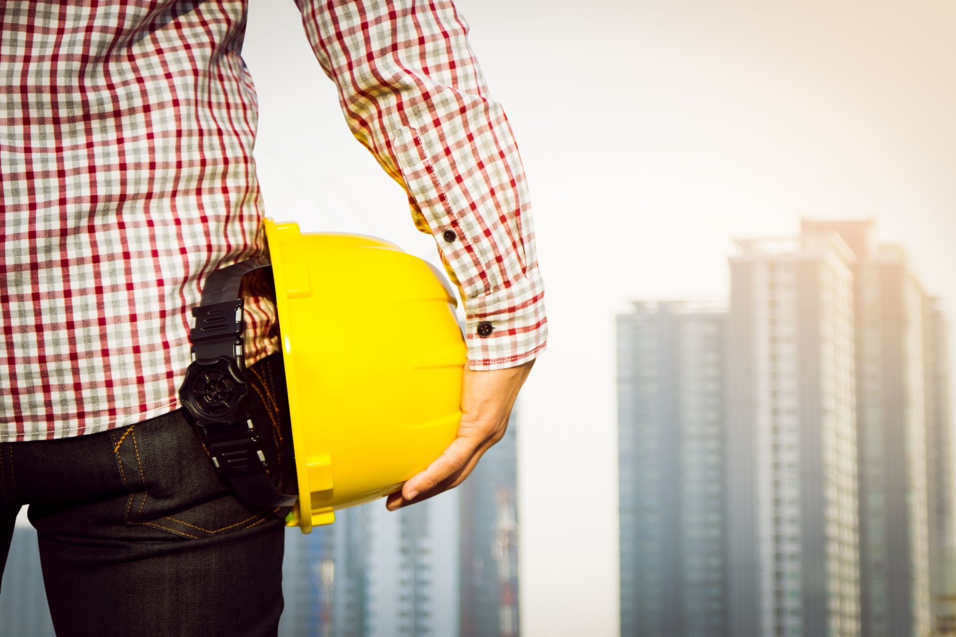 A construction worker is holding a yellow hard hat in front of a city.