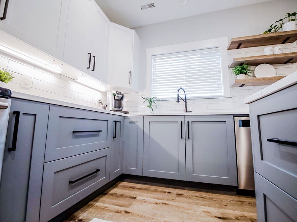 A kitchen with gray cabinets , white counter tops , a sink , and a window.