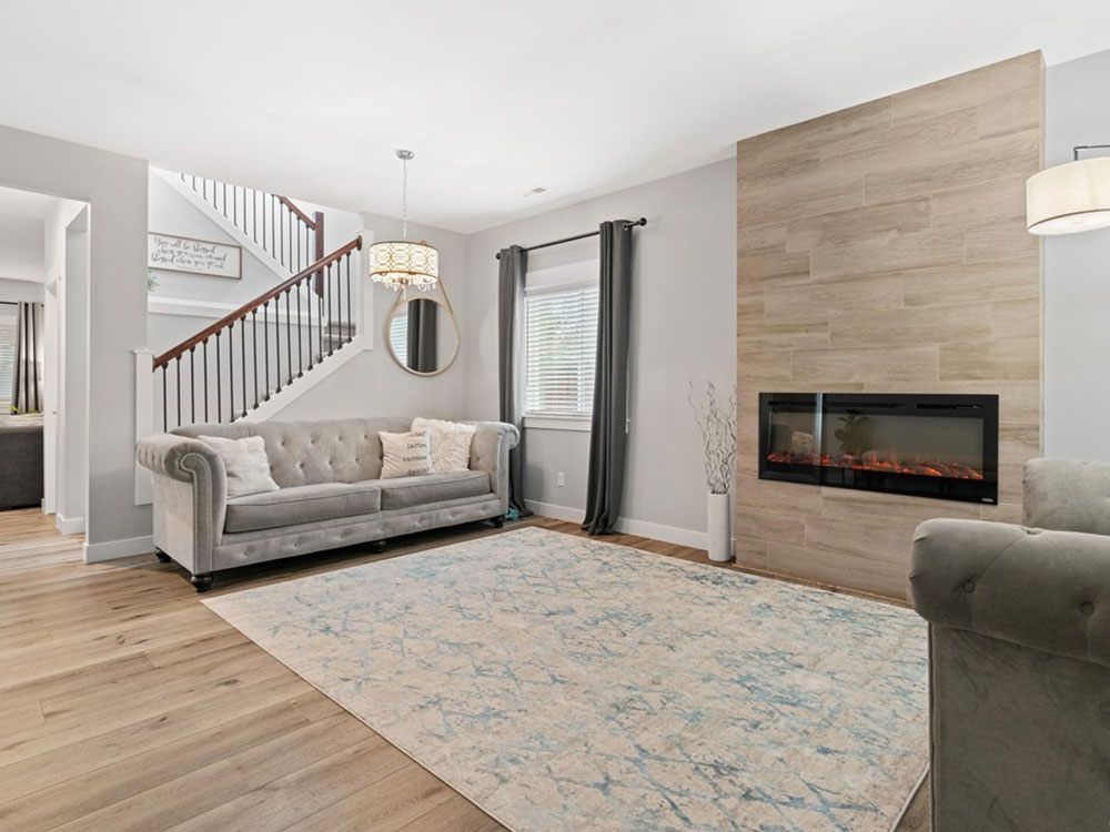 Bright living room with gray sofa, patterned rug, wood floors, staircase, and a fireplace wall