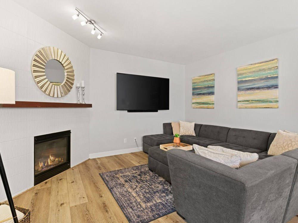 Modern living room with gray sectional, wall-mounted TV, fireplace, and colorful artwork on white walls