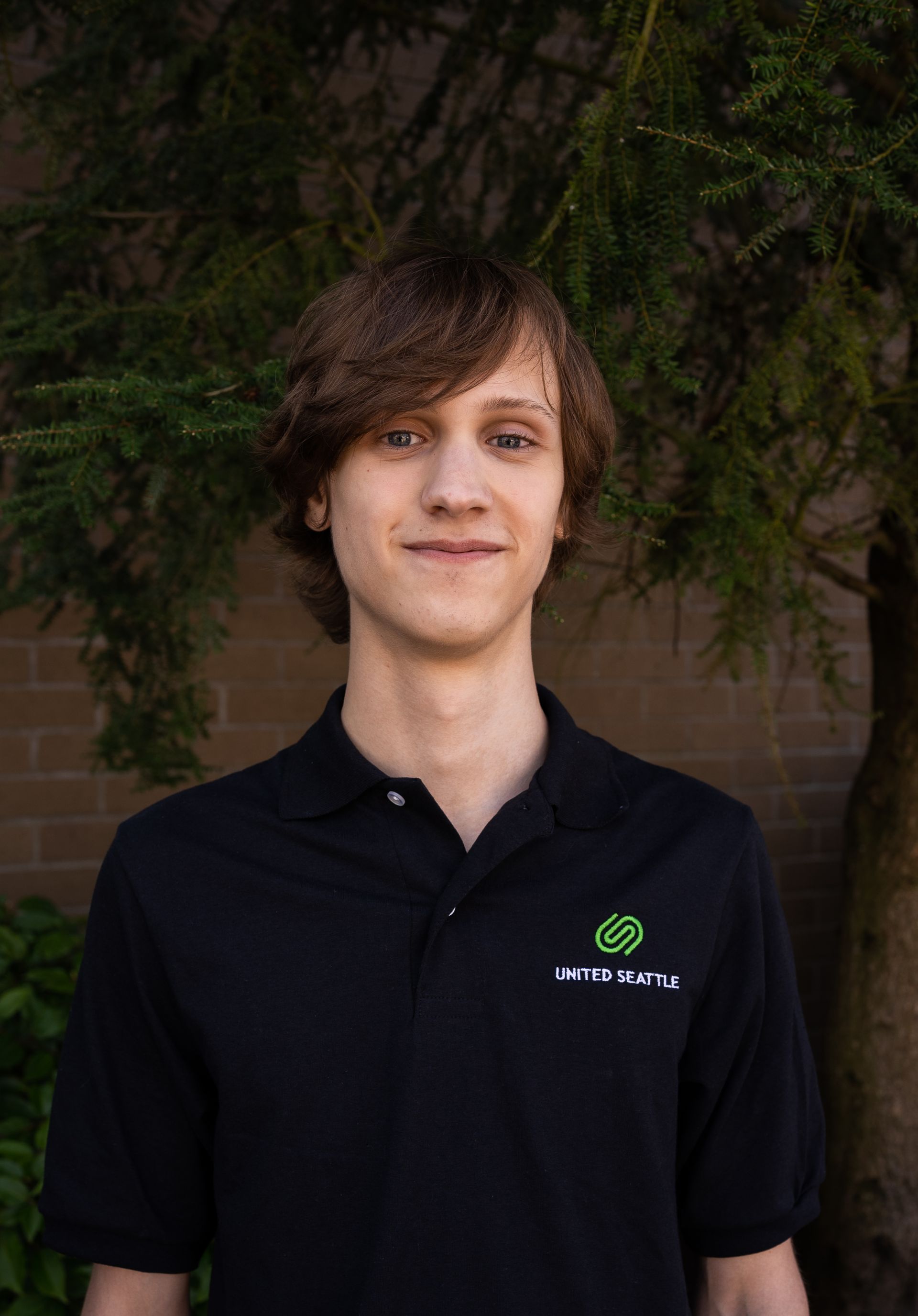 A young man wearing a black polo shirt is standing in front of a tree.