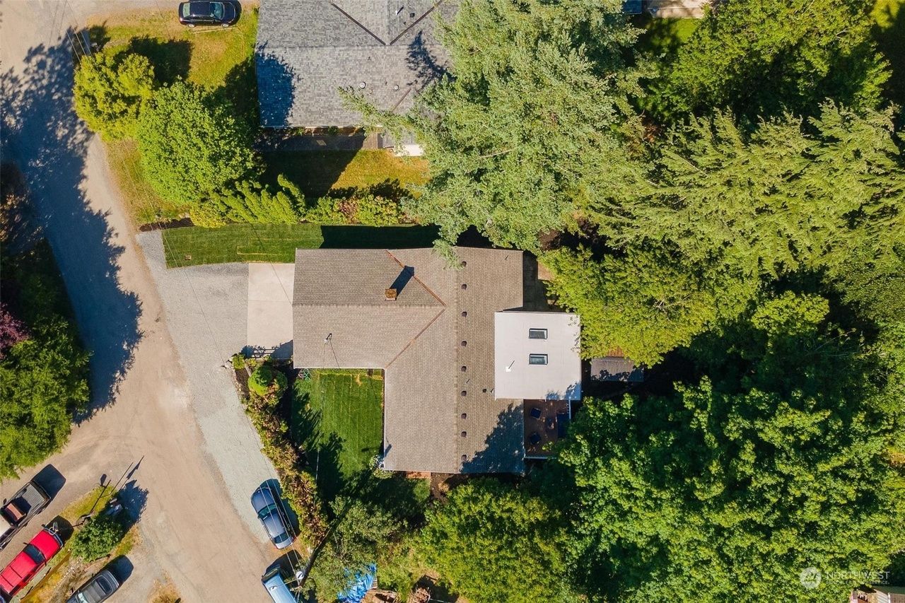 Aerial view of a house surrounded by dense trees, with a driveway and parked cars nearby.