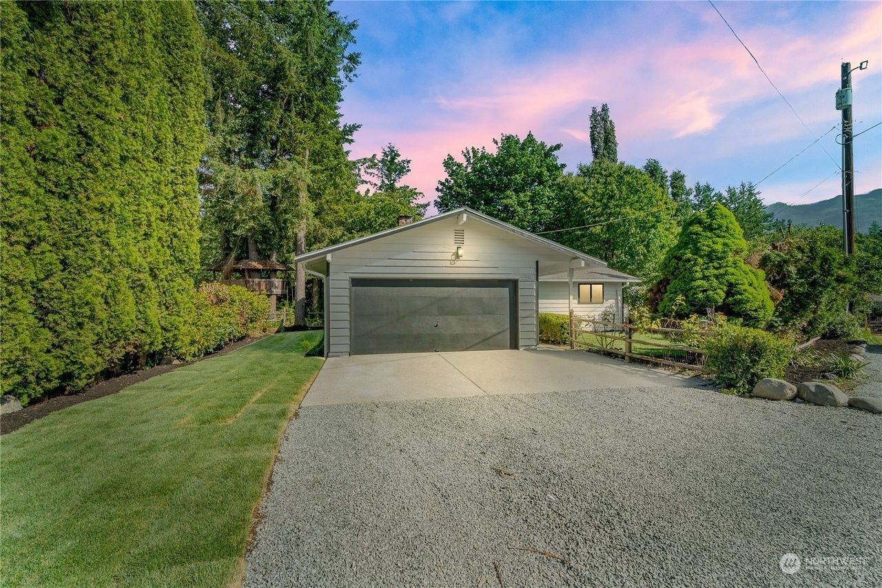 Driveway leading to a garage with a lit house front, surrounded by manicured lawn and trees at dusk.