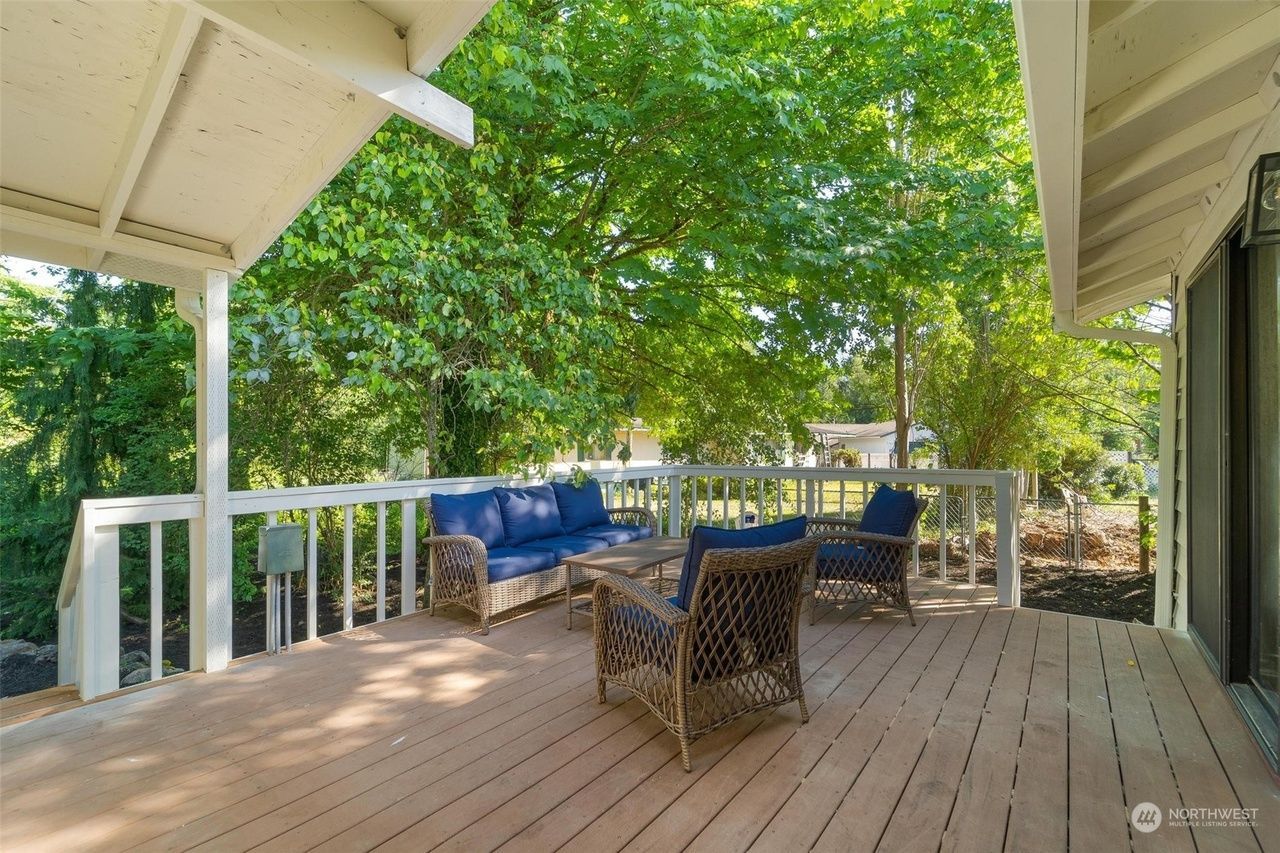 Covered wooden deck with blue cushioned chairs, wicker table, and trees beyond the railing