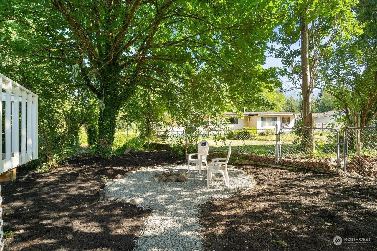 Gravel patio with white outdoor chairs under a large leafy tree in a shaded yard