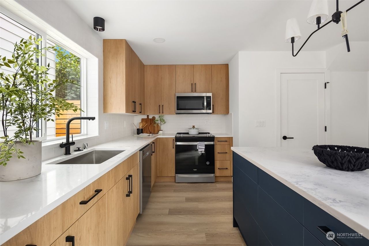 Bright modern kitchen with wood cabinets, white countertops, black fixtures, and a large window with greenery.