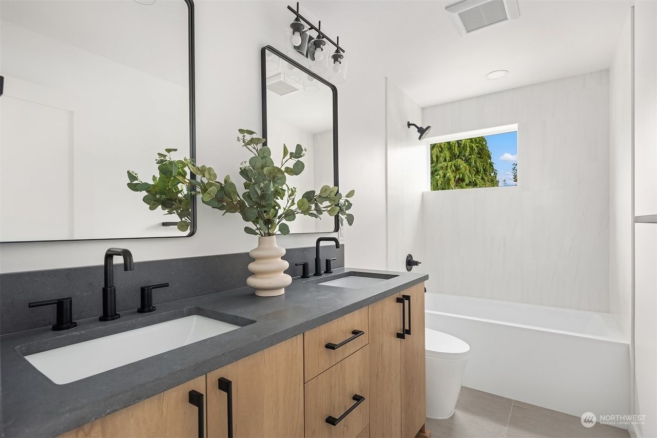 Modern bathroom vanity with black faucets, wood cabinets, plants, and a white bathtub by a window.