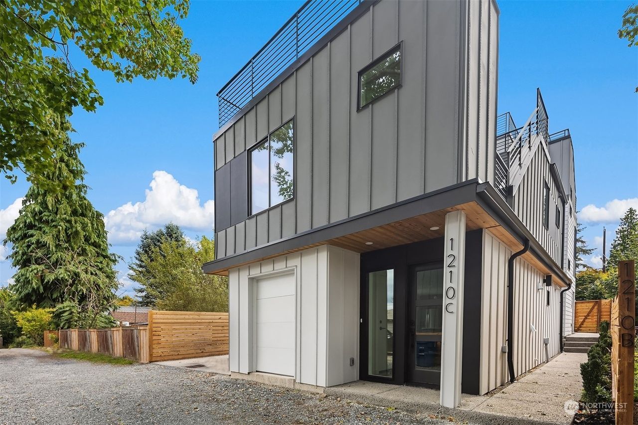 Modern gray two-story house with garage and large windows, viewed from a gravel driveway under a blue sky