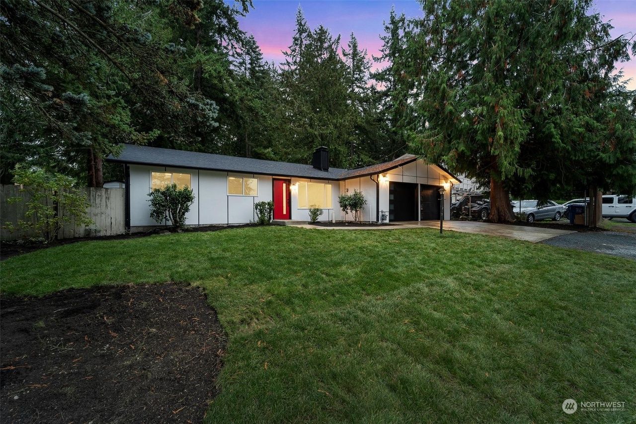 Single-story house at dusk with a red front door, lit porch, and a large grassy front yard surrounded by trees