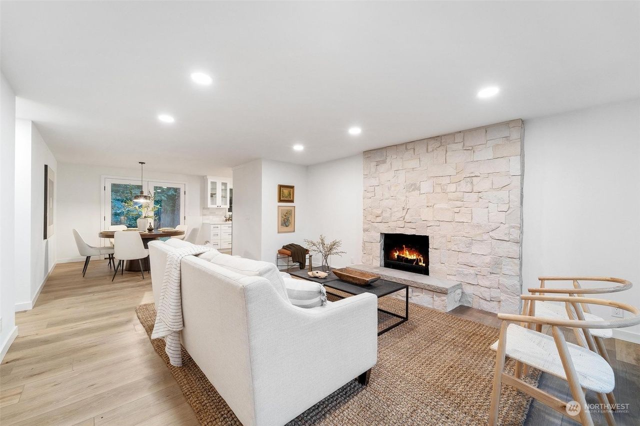 Bright modern living room with white sofa, stone fireplace, recessed lights, and dining area beyond
