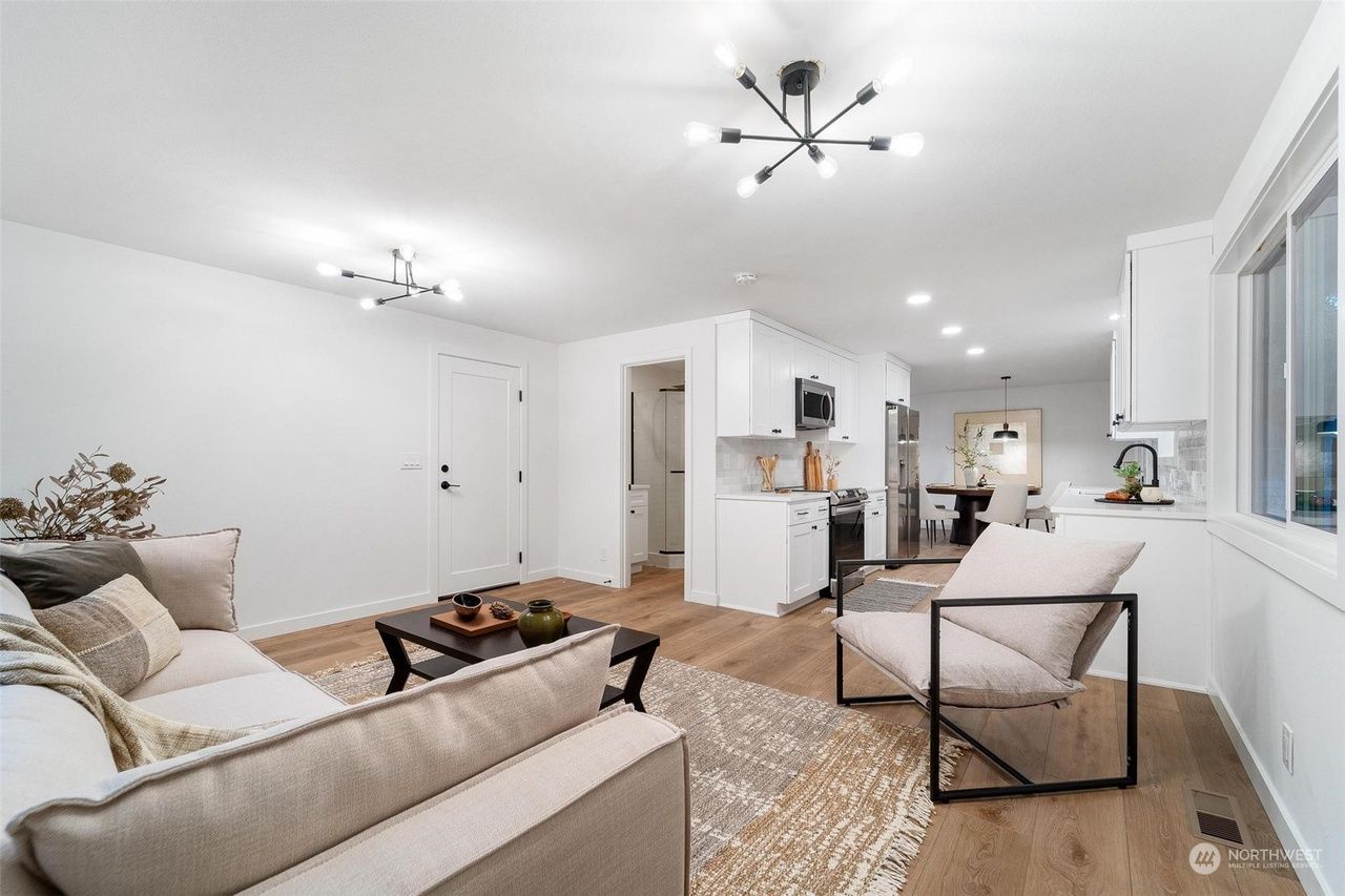 Bright open living room with white walls, beige sofas, rug, and modern ceiling lights leading to a kitchen.