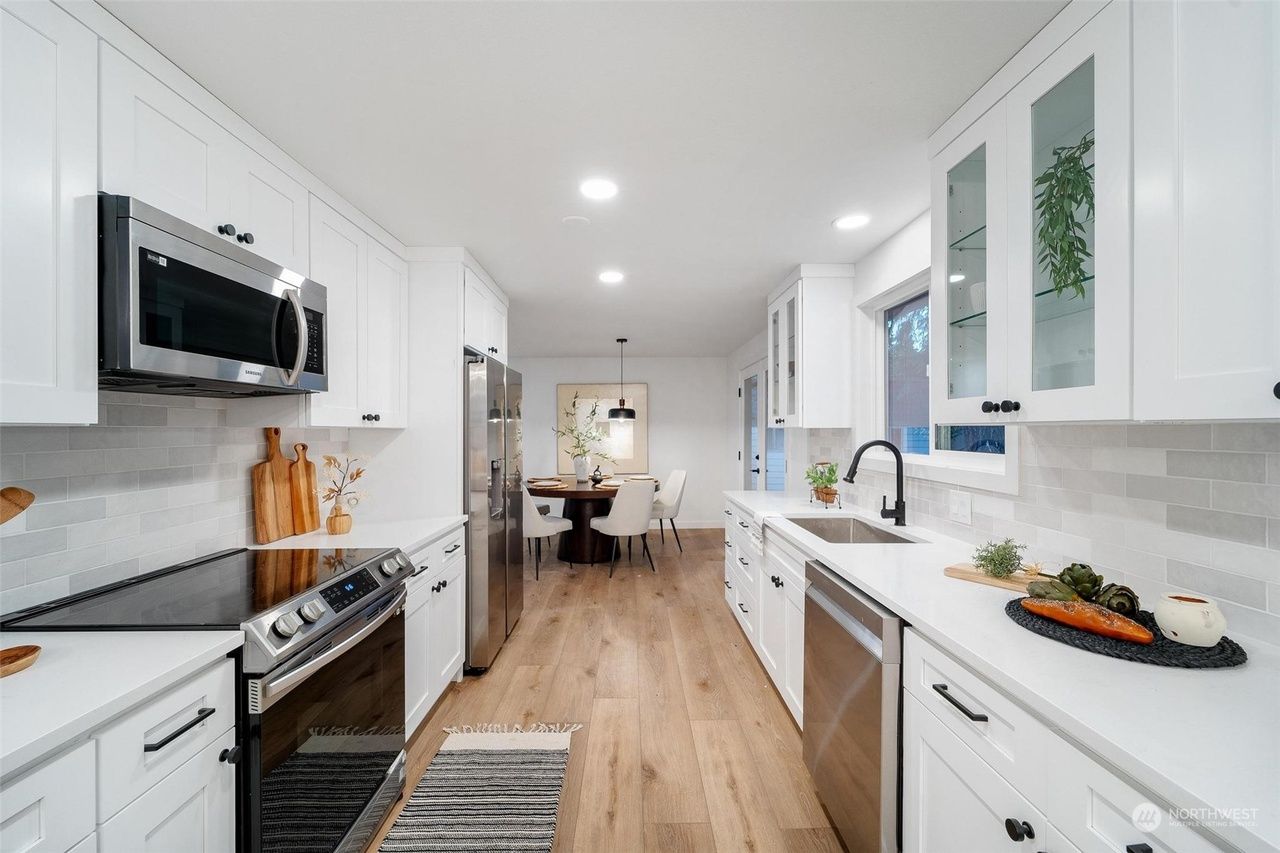 Bright modern white kitchen with stainless appliances, wood floor, and dining area beyond