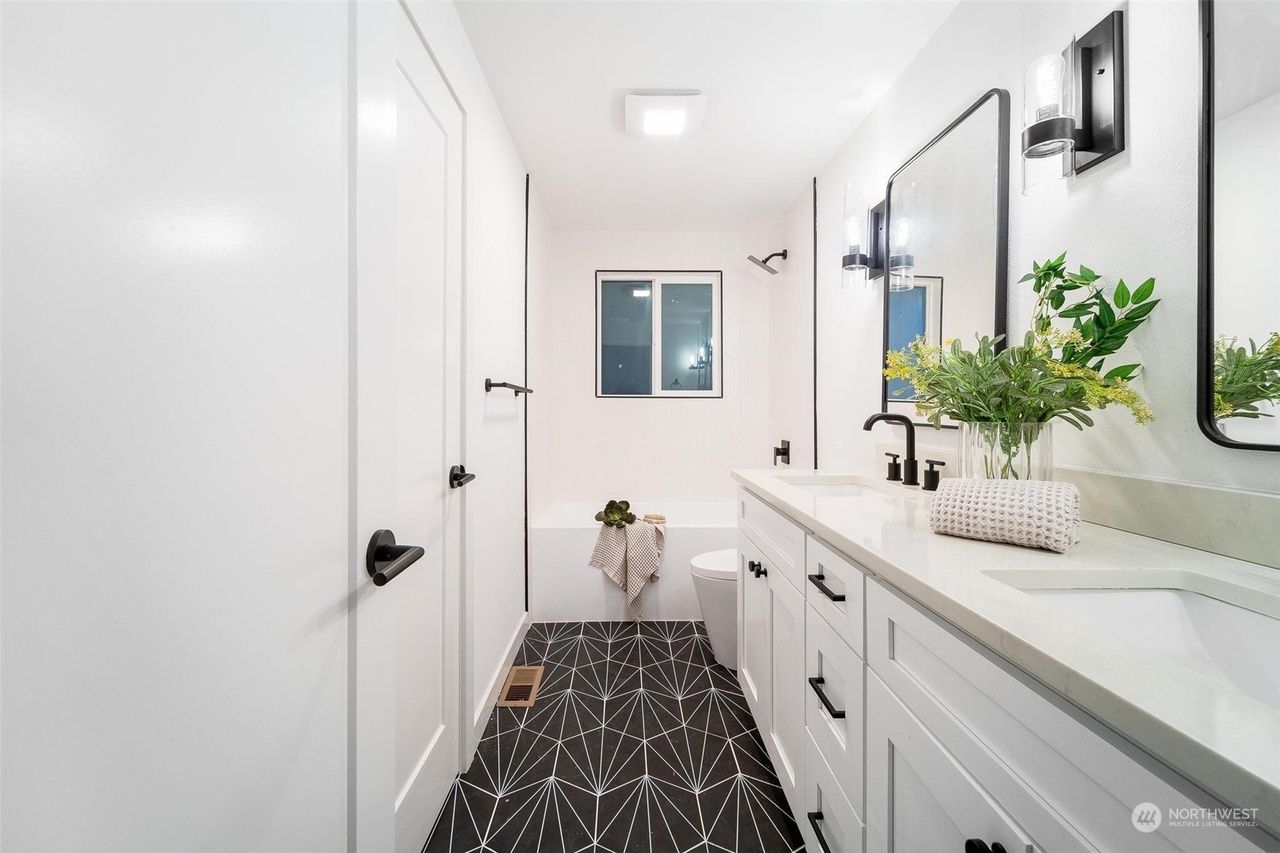 Bright white hallway with double doors, patterned rug, dresser with mirror, plants, and a window at the end.