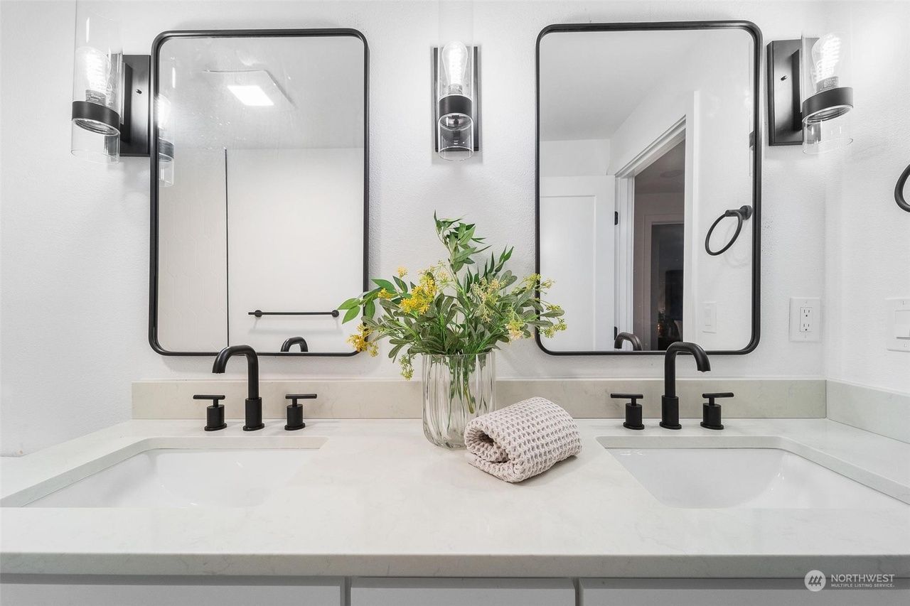 Bright bathroom vanity with two black faucets, framed mirrors, and a vase of flowers on the counter
