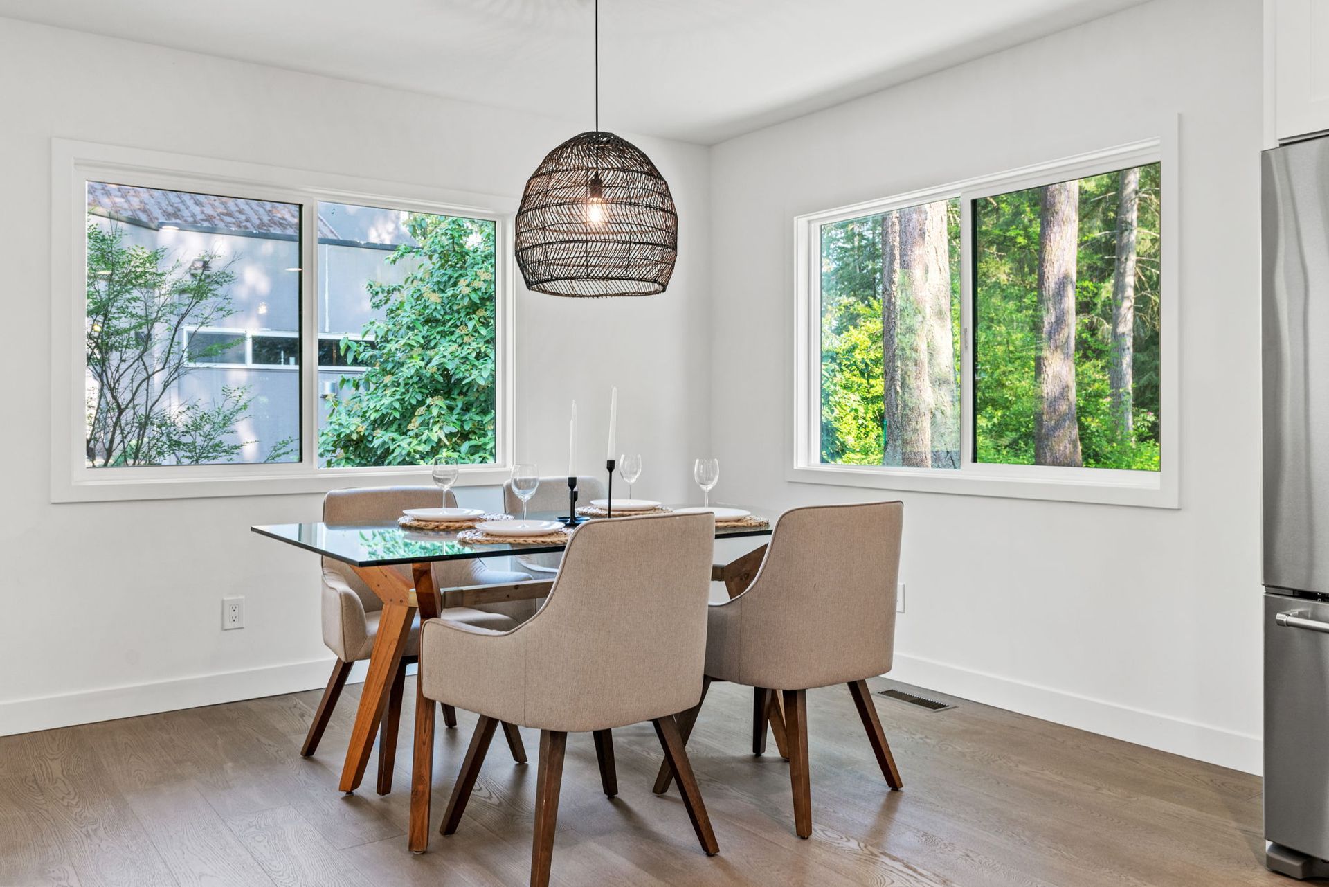 Bright dining room with a glass table, beige chairs, pendant light, and large windows overlooking greenery