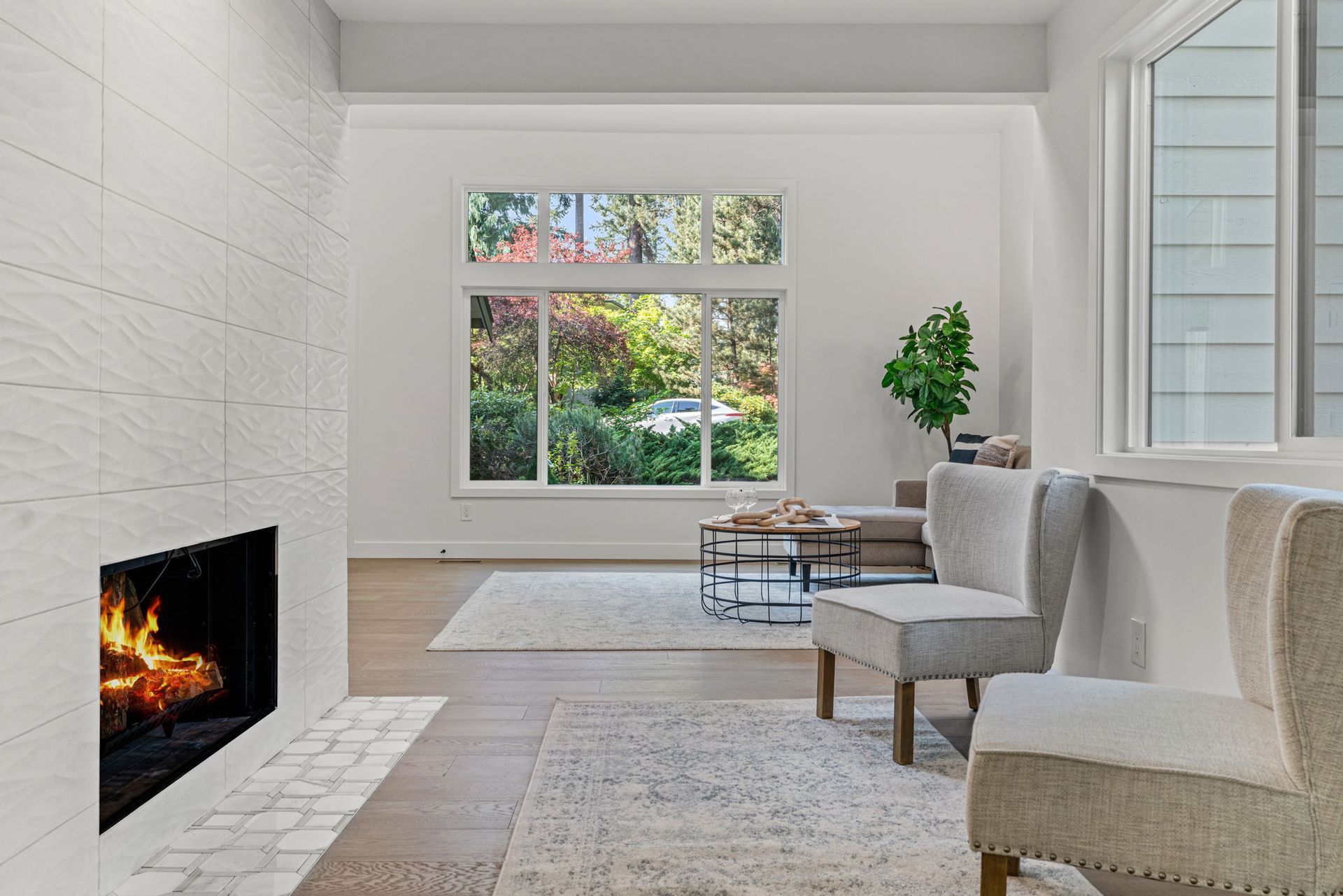 Modern living room with a fireplace, large window, gray chairs, and a view of greenery outside
