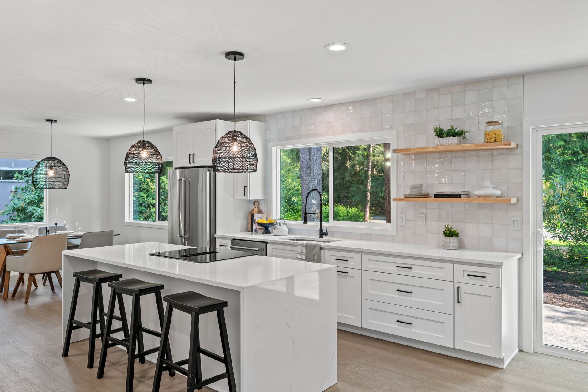 Bright modern kitchen with white island, black stools, pendant lights, and large windows.