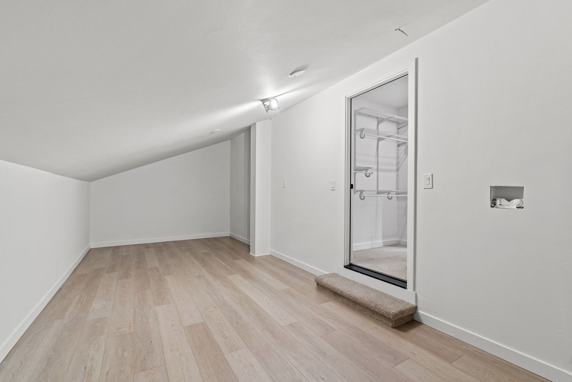 Empty attic room with sloped white ceiling, light wood floor, and open doorway to a tiled bathroom