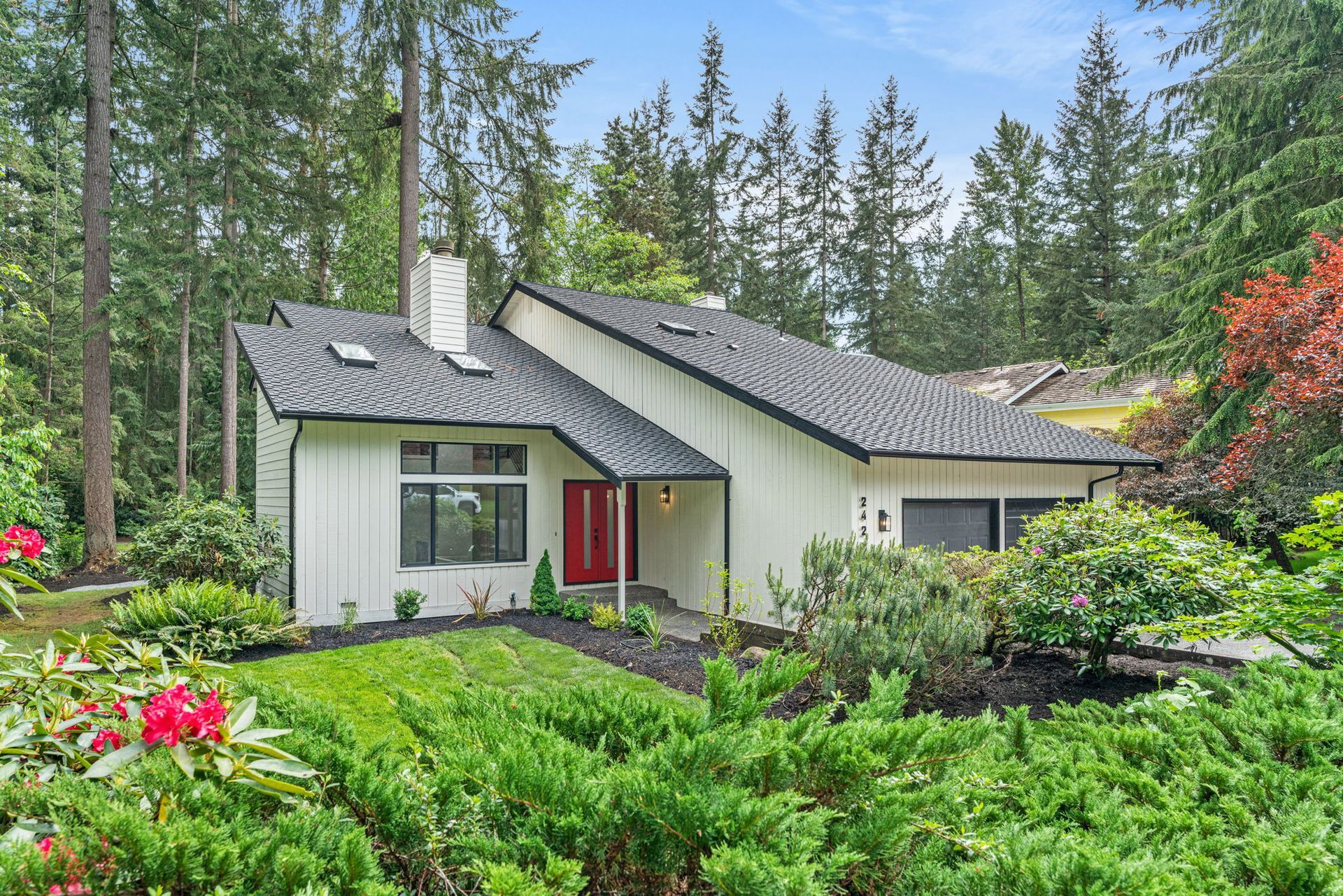 Modern house with gray roof and red door, surrounded by lush green garden and tall evergreen trees