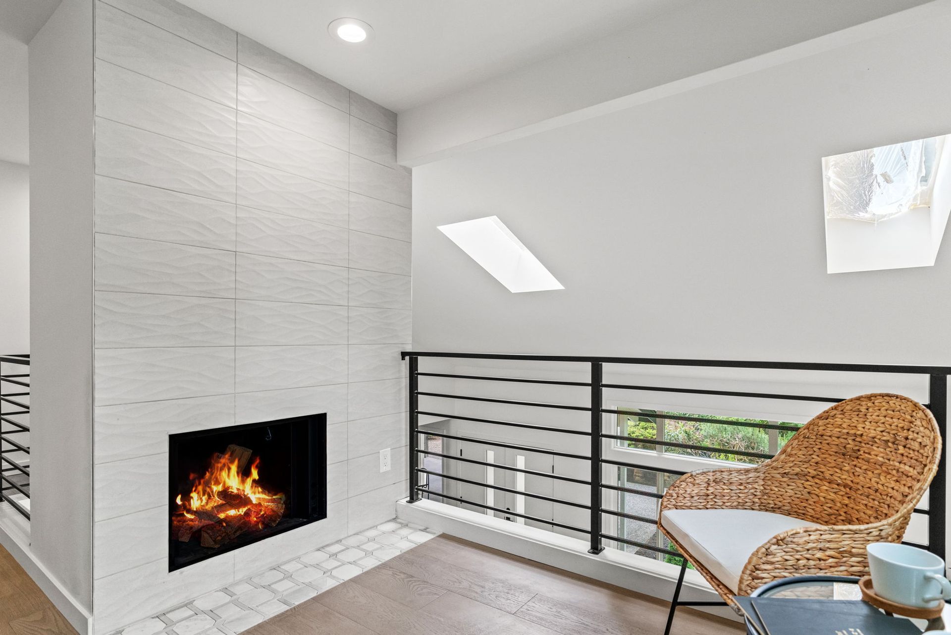 Modern living room with a white fireplace, balcony railing, skylights, and wicker chair by the window.