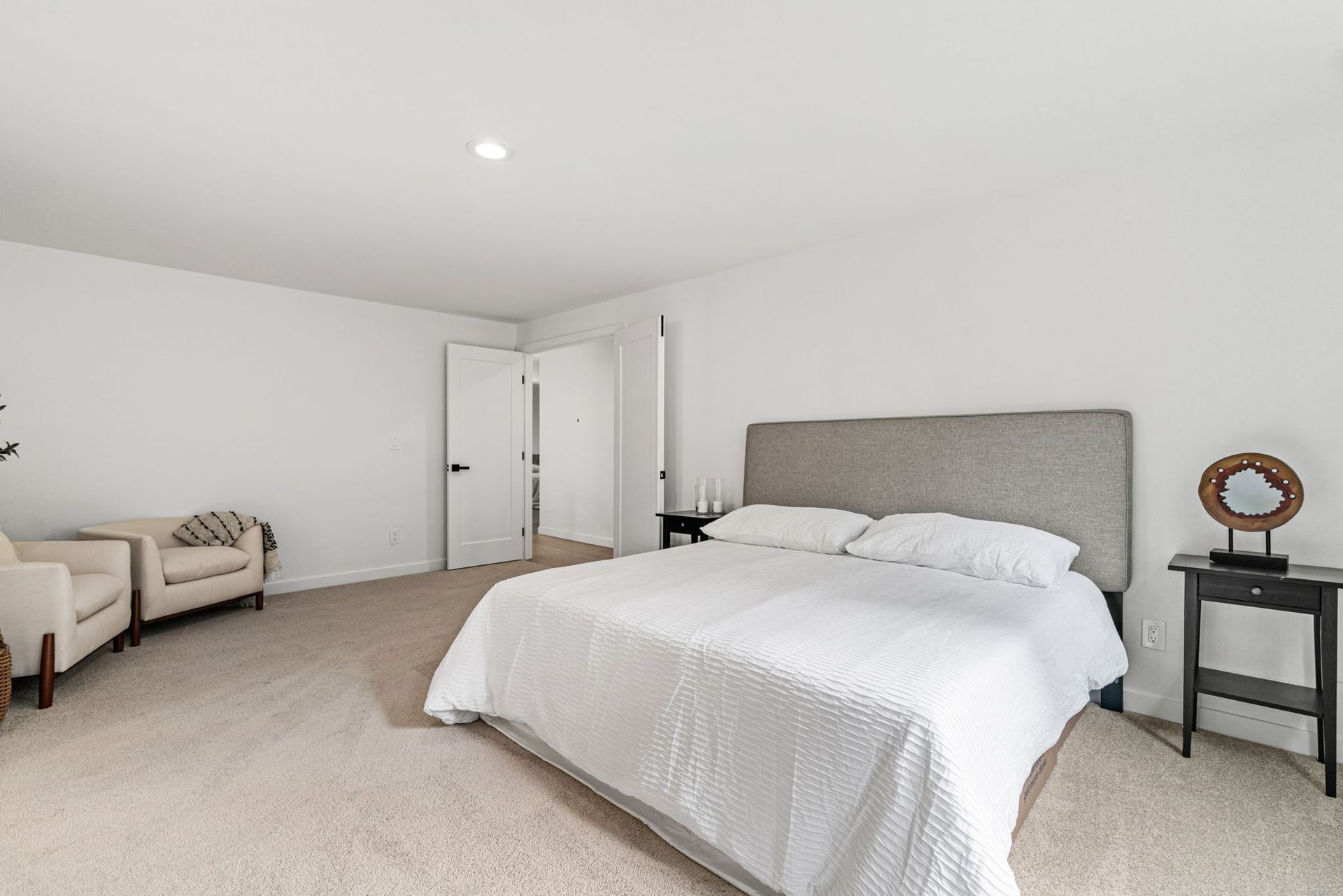 Bright, minimal bedroom with a white bed, beige carpet, and a chair by the wall.