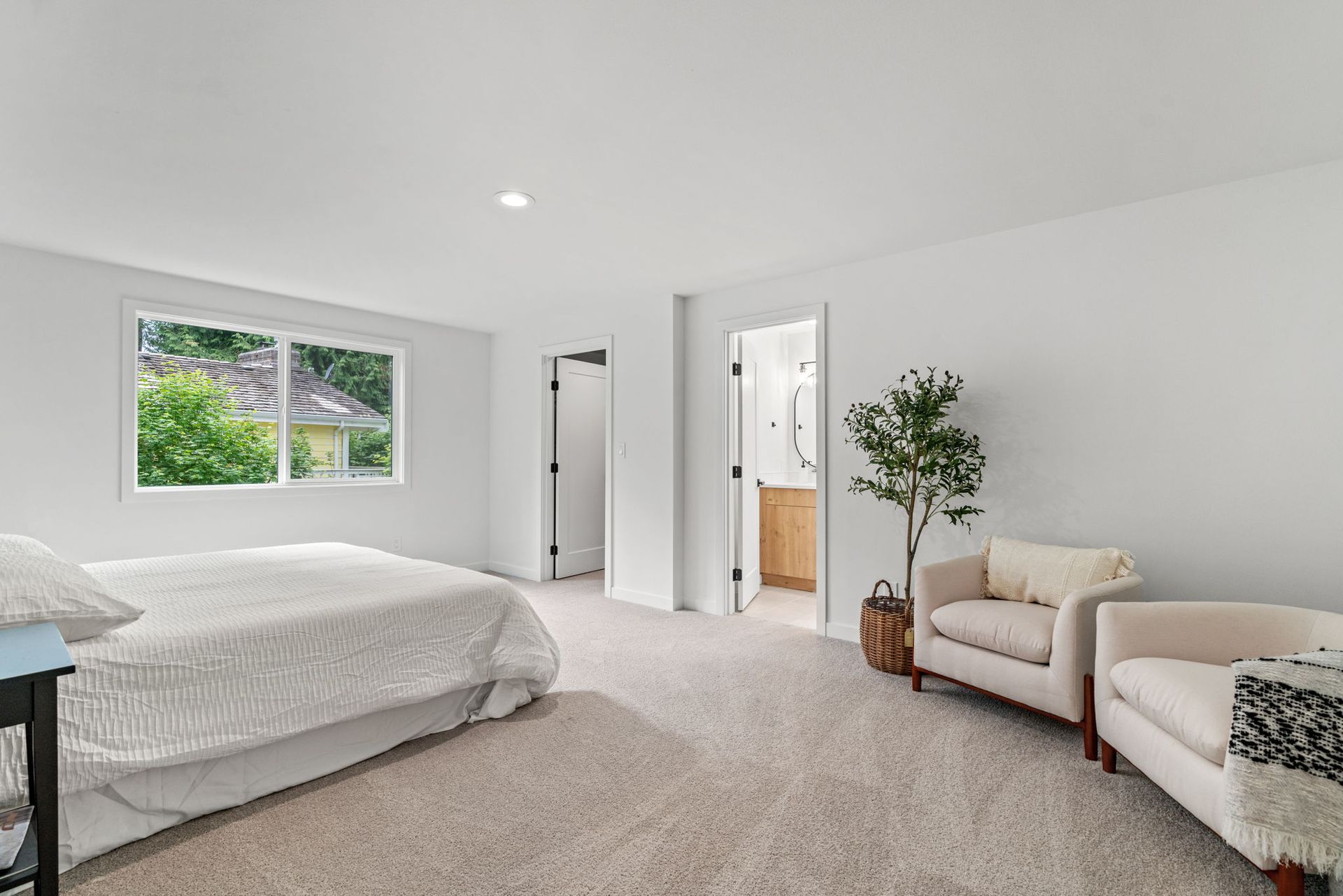 Bright bedroom with a bed, beige chair, potted tree, and window overlooking greenery