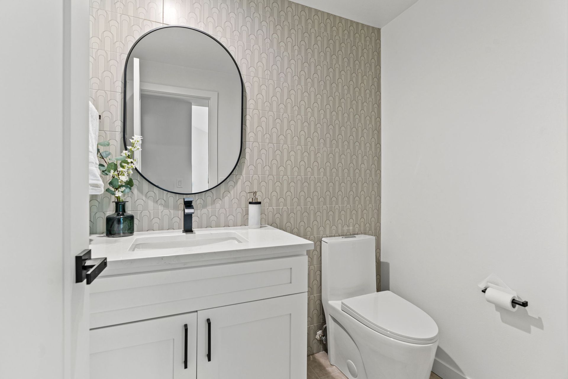 Small bathroom with white vanity, round mirror, and toilet against patterned wallpaper