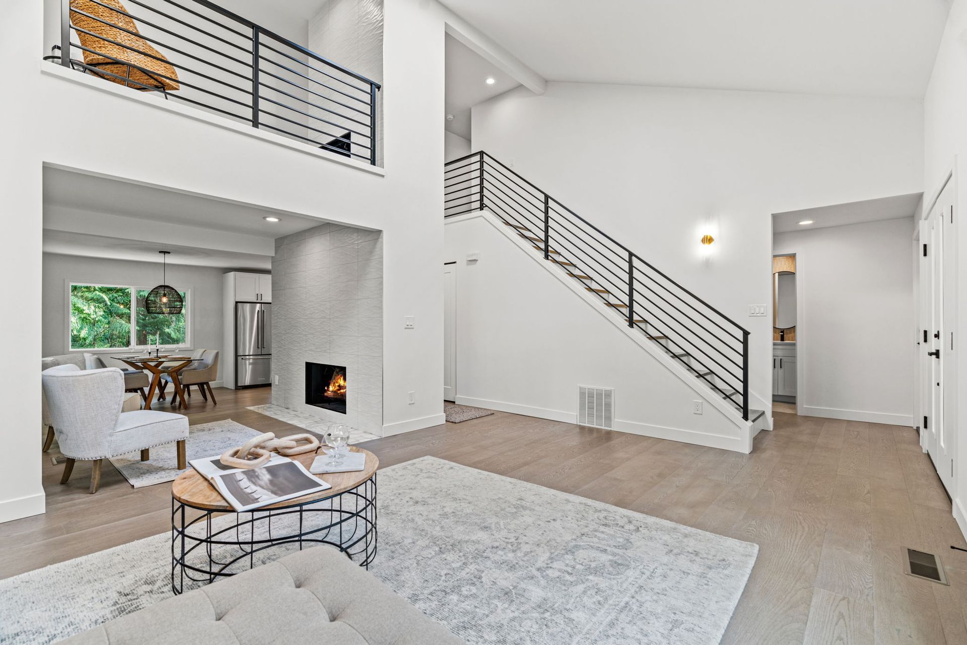 Bright modern living room with vaulted ceiling, white walls, staircase, fireplace, and large windows