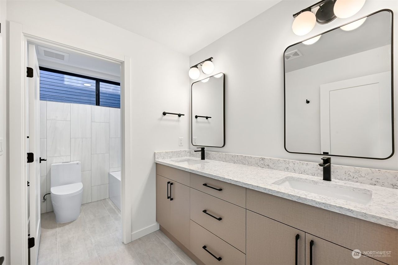 Bright modern bathroom with double vanity, mirrors, and a toilet in a tiled shower area.