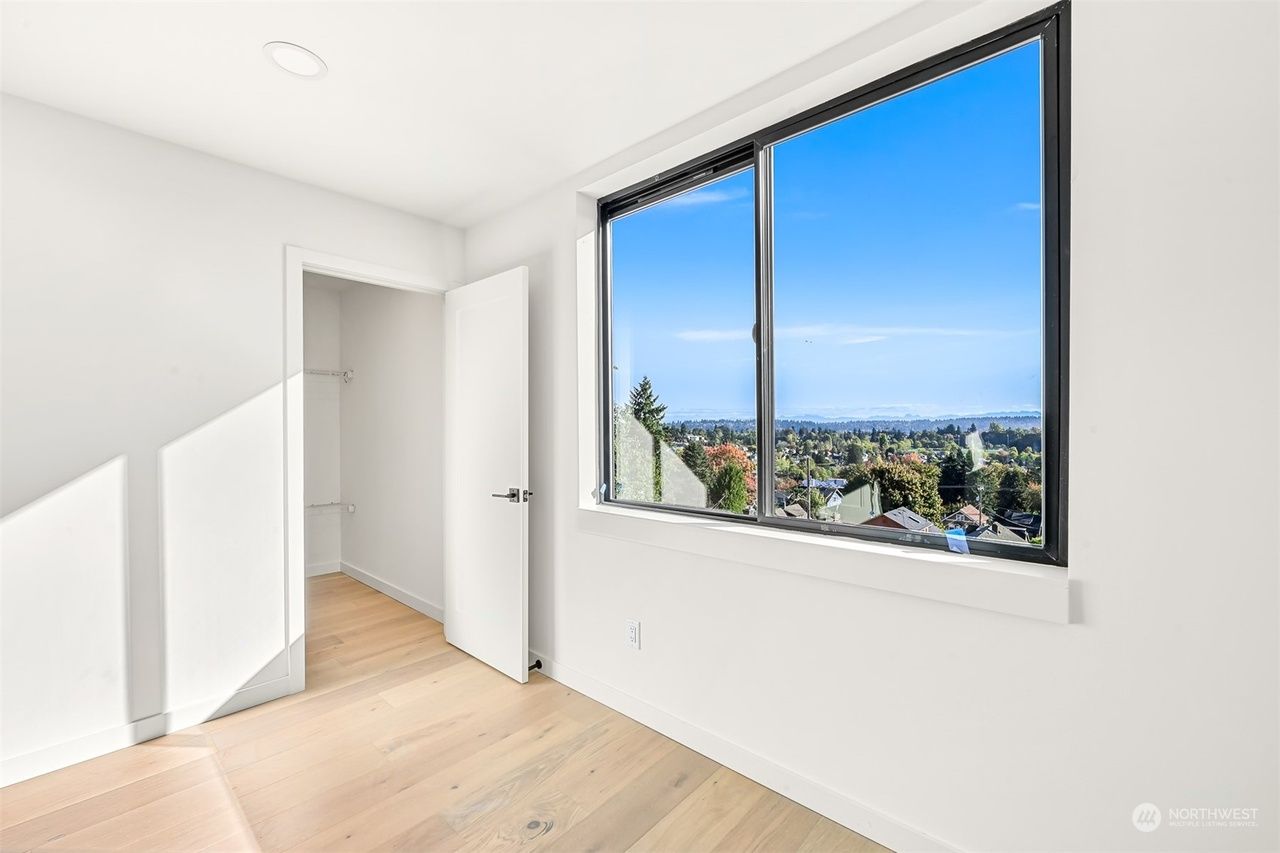 Bright empty white room with wood floors, open door, and large window overlooking a city view