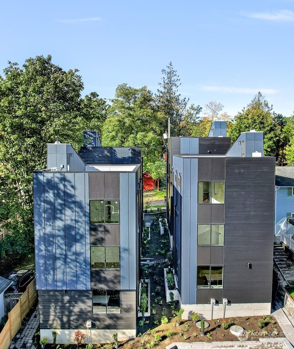 Two modern apartment buildings with glass balconies and rooftop structures among green trees under a blue sky