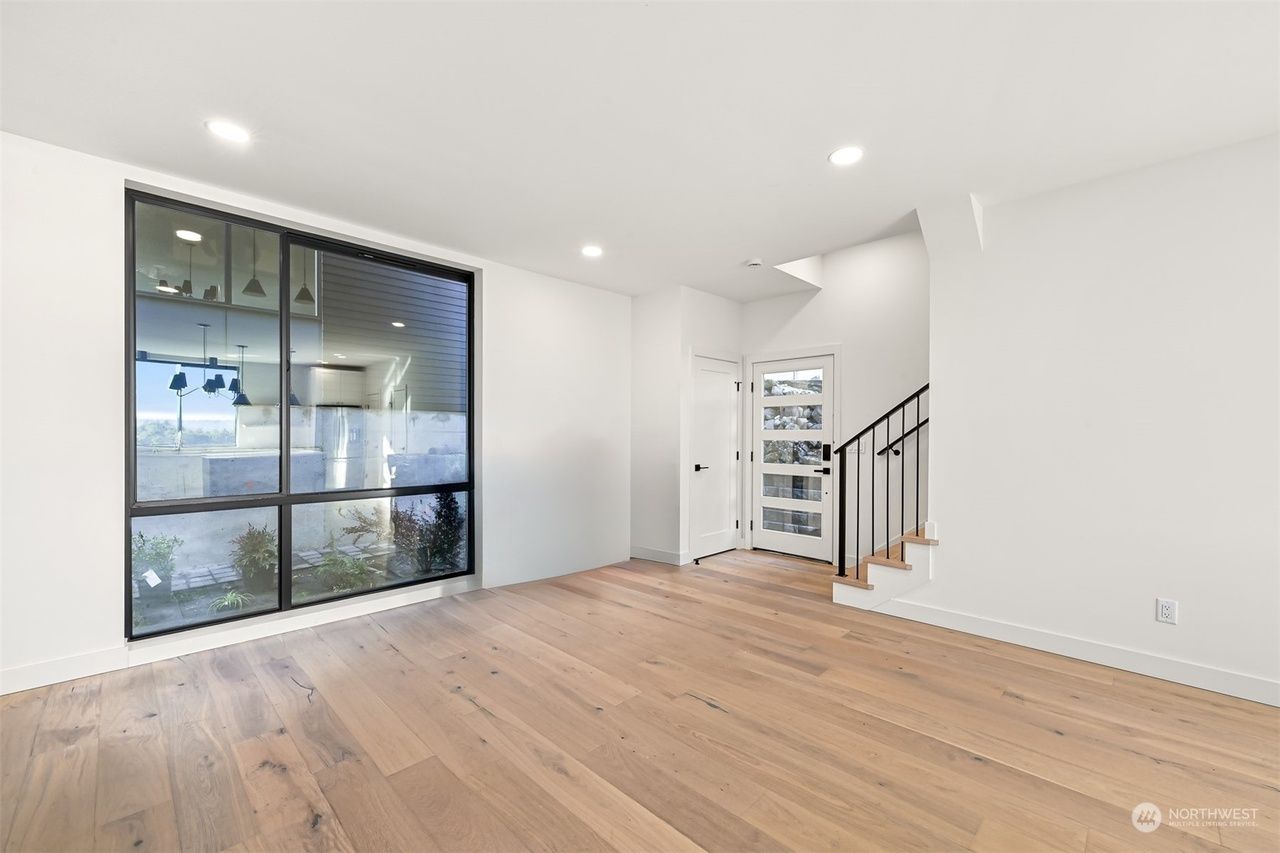 Bright empty living room with wood floors, large window, and staircase with black railing