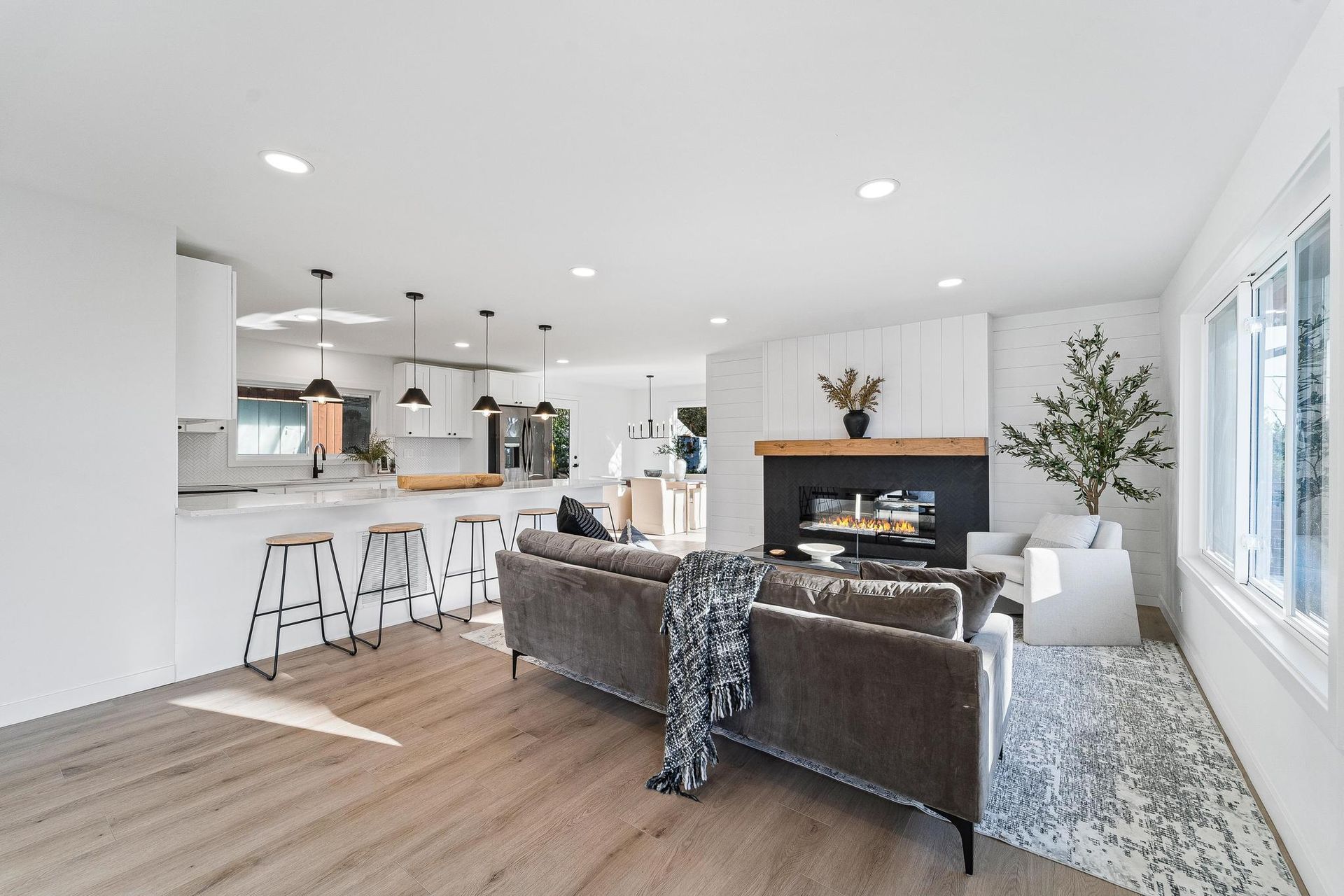 Bright open-concept living room with kitchen bar stools, gray sofa, fireplace, and large windows