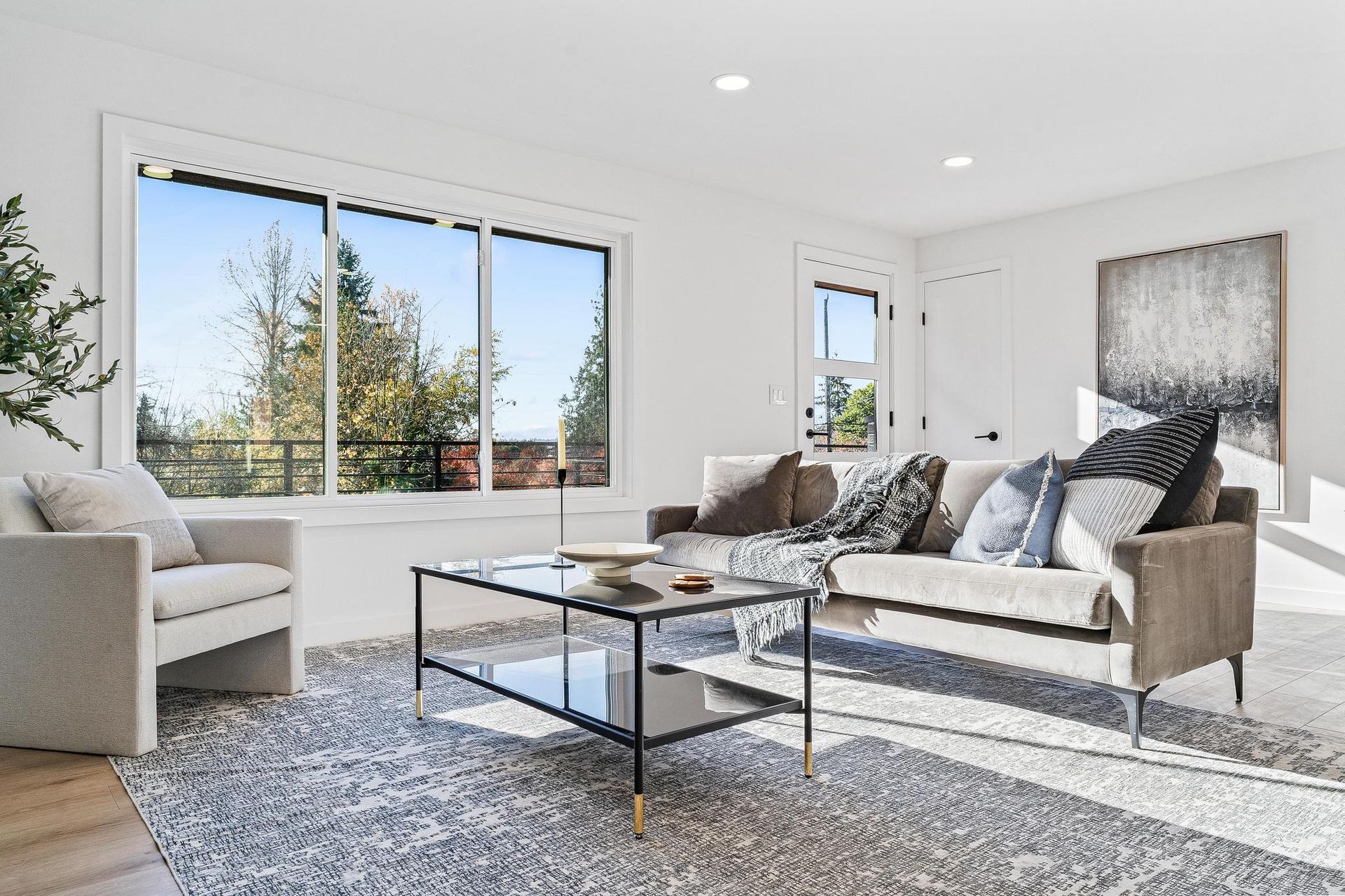 Bright modern living room with large windows, beige sofa, armchair, and glass coffee table on a shag rug.