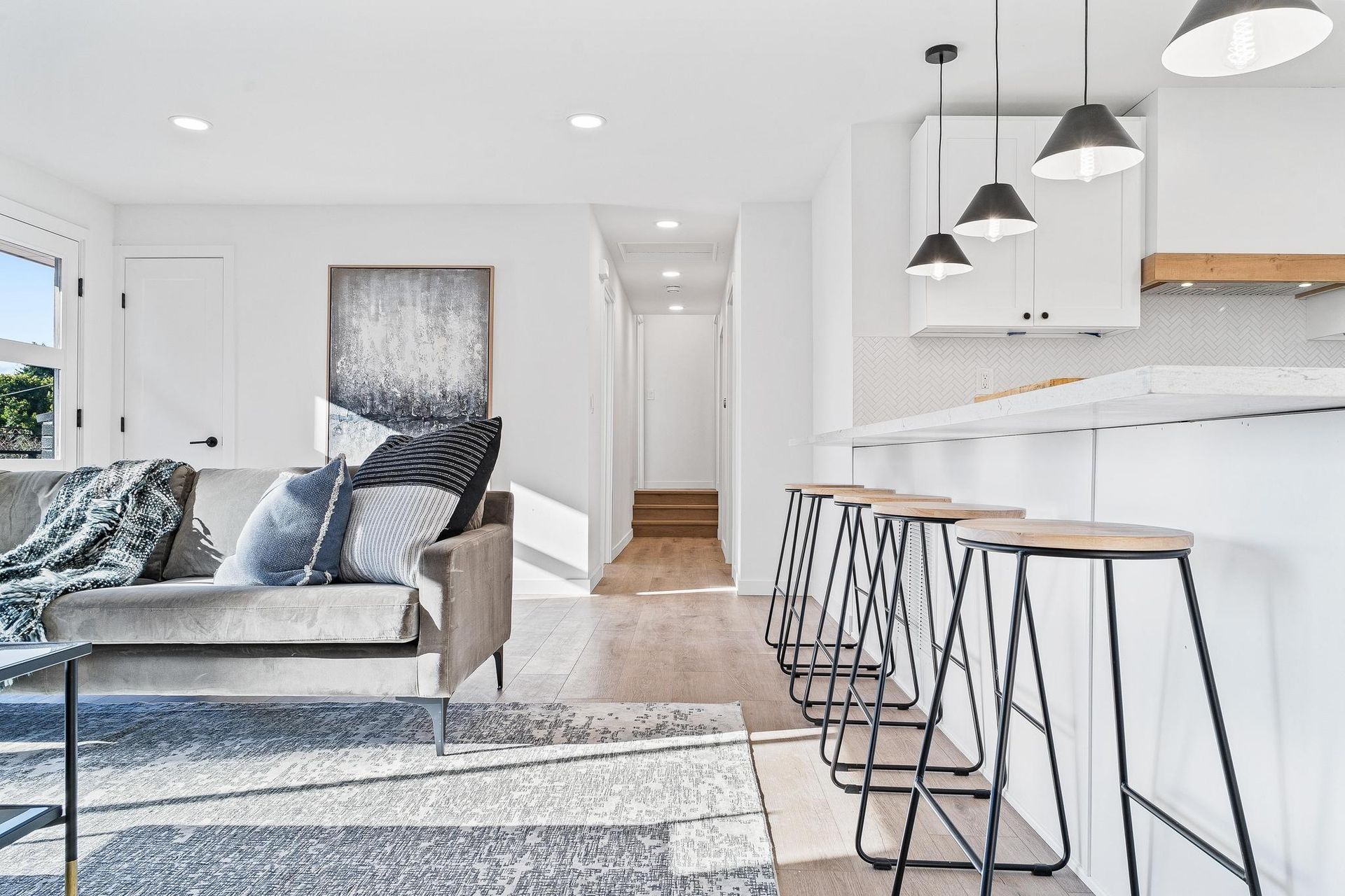 Modern white kitchen and living area with gray sectional sofa, bar stools, and pendant lights