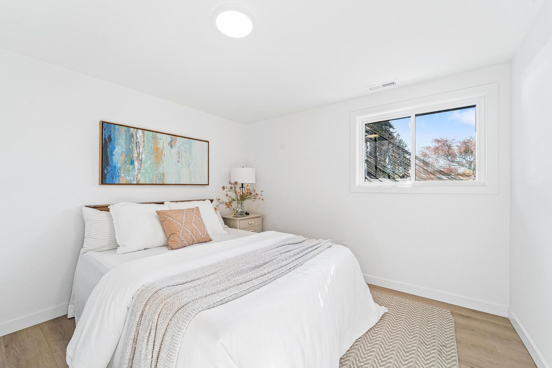 Bright white bedroom with a neatly made bed, abstract wall art, lamp, and window view