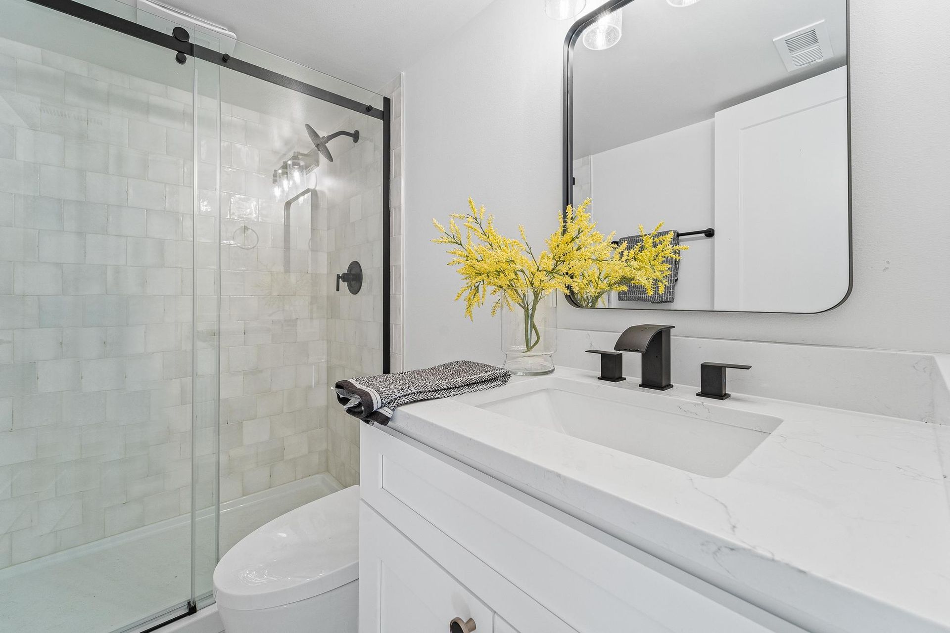 Bright modern bathroom with white vanity, mirror, shower, and yellow flowers.